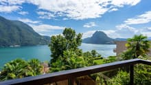 The image shows a scenic view of a lake surrounded by mountains and lush greenery. There is a balcony or terrace in the foreground, with palm trees and other tropical vegetation visible. The sky is blue with fluffy white clouds, creating a picturesque and