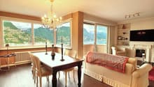 Spacious open-plan living and dining area with large windows offering a scenic view of the mountains and lake. The room features a wooden dining table with chairs, a plush sofa, and a decorative chandelier. The decor has a warm, cozy feel with neutral ton