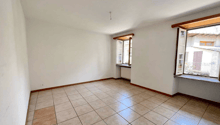 This image shows a spacious room with white walls and a tiled floor. The room has large windows that provide natural light and a view of the outdoors. The room appears to be empty, with no furniture or decor visible.