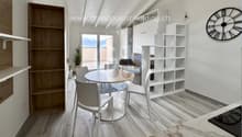 A room with white and gray furniture, including a desk and chairs, a round table, shelves, and a clock on the wall. There is a glass door leading to a balcony with a view of the outdoors.
