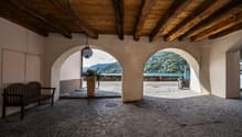 open area with wooden beams ceiling, cobblestone floor, wooden bench, potted plant, two arched openings leading to outside view