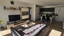 Gray couch, wooden floor, TV mounted on wall, dining table with chairs, kitchen island with black chairs, white walls, potted plant