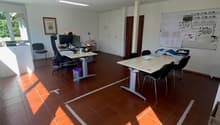 Large open office space with hardwood floors, white walls, and modern furniture including desks, chairs, and a whiteboard. The room has good natural lighting from the windows.