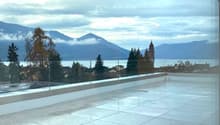 The image shows a balcony or terrace overlooking a scenic landscape. In the distance, there are mountains and a church tower visible. The balcony has a tiled floor and glass railings, providing an unobstructed view of the surrounding area.