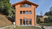 Two-story house, bright orange exterior, multiple large windows, black railing on balcony, located on a hillside