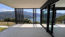 Large glass windows and sliding doors overlooking a scenic lake and mountain landscape, with a spacious outdoor terrace or balcony area.