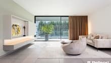 Spacious living room with large windows offering a view of the surrounding greenery. The room features a modern and minimalist design, with a sleek TV console, a comfortable sofa, and a stylish armchair. The curtains add a touch of warmth to the space.