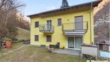 2-story yellow house with balconies, surrounded by trees and a grassy yard, with a rocky cliff in the background