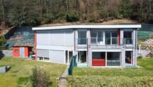 Two-story modern house with a flat roof, large windows, and a garage. The exterior is made of a combination of white siding and stone. There is a small paved area in front of the house that could be used for parking. The house is surrounded by a grassy ya