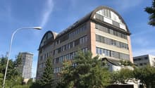 A multi-story office building with a curved, arched roof and large windows. The building has a parking area in front and is surrounded by trees and other buildings.
