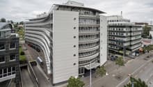 Large multi-story commercial building with a curved facade, glass windows, and a parking area in front. The building appears to be located in an urban setting with other nearby buildings and infrastructure visible.