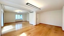 This image shows a spacious, open-plan room with hardwood floors and large windows. The room has a modern, minimalist design with a long, rectangular light fixture hanging from the ceiling. The walls are painted a neutral color, and there is a door visibl