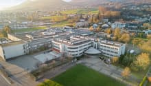 The image shows a large multi-story building complex surrounded by a grassy area and trees. The buildings have a modern, industrial design with flat roofs and large windows. The complex appears to be situated in a hilly, mountainous area with a town visib