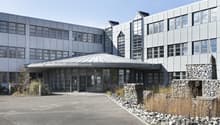 Large modern commercial building with a glass and metal facade, a domed roof structure, and a paved parking area in the foreground. The building has multiple floors and large windows. There is also a gabion wall feature with stones near the entrance.