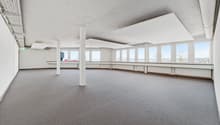 Large open-plan office space with high ceilings, white walls, and large windows providing natural light. The room has a carpeted floor and several support columns. The space appears to be unoccupied and ready for use.