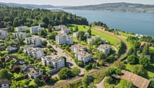 Aerial view of an urban residential area near a lake and mountains, multiple buildings with solar panels, some with balconies, surrounded by greenery.