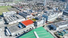 Large industrial area with multiple buildings, some with red tennis courts, multiple solar panels, extensive parking spaces, and multiple cranes and construction vehicles