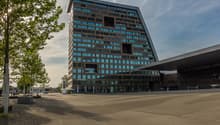 A large, modern office building with a glass and steel facade. The building has many floors and windows, and is surrounded by trees and a paved plaza area in front of the entrance.