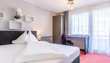 Spacious bedroom with a large bed, white bedding, and a desk area. The room has large windows with curtains, providing natural light. There are shelves on the wall and a decorative plant. The room has a modern and cozy feel.