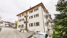 3-story apartment building with white exterior, green shutters, and wooden roof. The building has several balconies and a staircase leading up to the entrance. There is a car parked in front of the building, and the surrounding area is lush with trees.