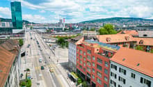 The image shows a panoramic view of a city skyline. There are tall modern buildings, including a green glass skyscraper, as well as older red-roofed buildings. The city is surrounded by hills and mountains in the background. The streets are busy with traf