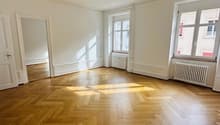 An empty room with parquet flooring, white walls, three windows, and two radiators. One of the rooms has a mirror.