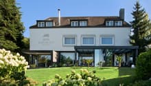 4-star Hotel Burgblick, 3-story building with a brown tiled roof, large windows, and a glass-enclosed patio area. The hotel is surrounded by a well-manicured lawn and lush landscaping, including flowering plants and shrubs.