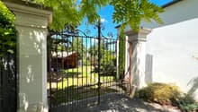 Open gate, wide gravel pathway, garden with various plants, trees, and a shed in the background