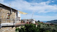 Stone wall building, terrace with awning, potted plants, outdoor table and chairs, scenic view of the ocean and hills