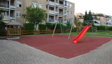 a playground with a slide, a swing set, and a picnic bench, a small wooden house, a paved ground, and a green grass area with trees