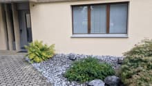 paved driveway, balcony with metal railing, beige wall, gray door, small plants, gravel and rocks
