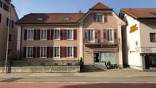 A multi-story building with a red tile roof, pink exterior walls, and white window frames. The building has a balcony on the upper floors and a storefront on the ground level.