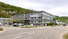 A multi-story commercial building with a gray exterior and large windows. The building appears to be located in a hilly, forested area with other smaller buildings and vehicles visible in the surrounding area.
