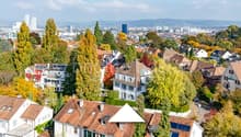 Multiple buildings in urban setting, brown roofs, green trees, cityscape view, mountains in background.