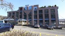 Large office building with many windows, roof terrace, large parking area in front of the building