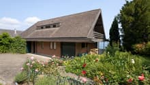 2-story brick house with a slanted roof, surrounded by a well-maintained garden with various flowers and plants. The house has a garage attached to the side.
