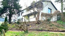 Two-story house with a balcony, surrounded by trees and vegetation. The house has a sloped roof and large windows. There is a raised garden area in front of the house.