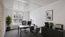 A modern office space with a desk, computer, and office chair. The room features a white wall, a radiator, and a window with a view of a building. There is a plant on the desk and a picture frame on the shelf. The room is empty and well-lit.