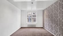 This image shows an empty office space with white walls, a large window, and a textured accent wall. The room has a modern, minimalist design with recessed lighting fixtures in the ceiling. The floor is covered in a neutral-colored carpet. Overall, the sp