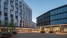 Modern multi-story office building with large glass windows, surrounded by a paved courtyard area with planters and seating spaces.