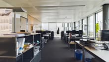 Open office space with cubicles, desks, and storage units. The ceiling has exposed beams and the walls are mostly glass, allowing natural light to enter the space. There are various office equipment and supplies visible, including a dishwasher.