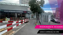 Facade of a commercial building with a sign reading 'ravaux intérieurs & extérieurs Tie', a closed shop with a canopy, a sidewalk, some barriers on the road, parked cars, trees and a building in the background.