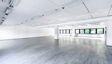 Large open office space with white walls, gray laminate flooring, and overhead fluorescent lighting. The space has large windows along one side providing natural light. The room appears to be empty and ready for use.