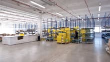 Large industrial warehouse space with shelving units, storage boxes, and a reception desk. The space has high ceilings, concrete floors, and overhead lighting.
