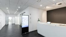 The image shows a modern office space with a reception area. The space has a clean, minimalist design with white walls, a gray floor, and a long white reception desk with a laptop on it. There is a large digital display screen in the center of the room, a