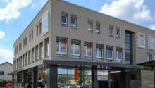 A 3 story building, grey walls, glass windows, signages, cars parked in front