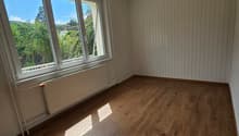 This image shows an empty room with hardwood flooring and white paneled walls. The room has large windows that provide a view of the surrounding greenery outside. The room appears to be a bedroom or living space with a simple and minimalist design.
