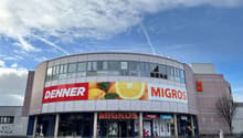 a modern, commercial building with storefronts, glass windows, and a curved facade. There are large advertisement boards with pictures of oranges on it.