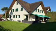 2-story house with a brown tiled roof, white exterior walls, and green shutters. The house has a covered patio or balcony area with a retractable awning. There is a garage and additional parking space on the property. The house is surrounded by a well-man