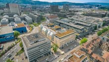 The image shows an aerial view of a dense urban cityscape with a mix of modern high-rise buildings and older structures. The city appears to be located in a hilly or mountainous area, with mountains visible in the background. The streets are lined with pa